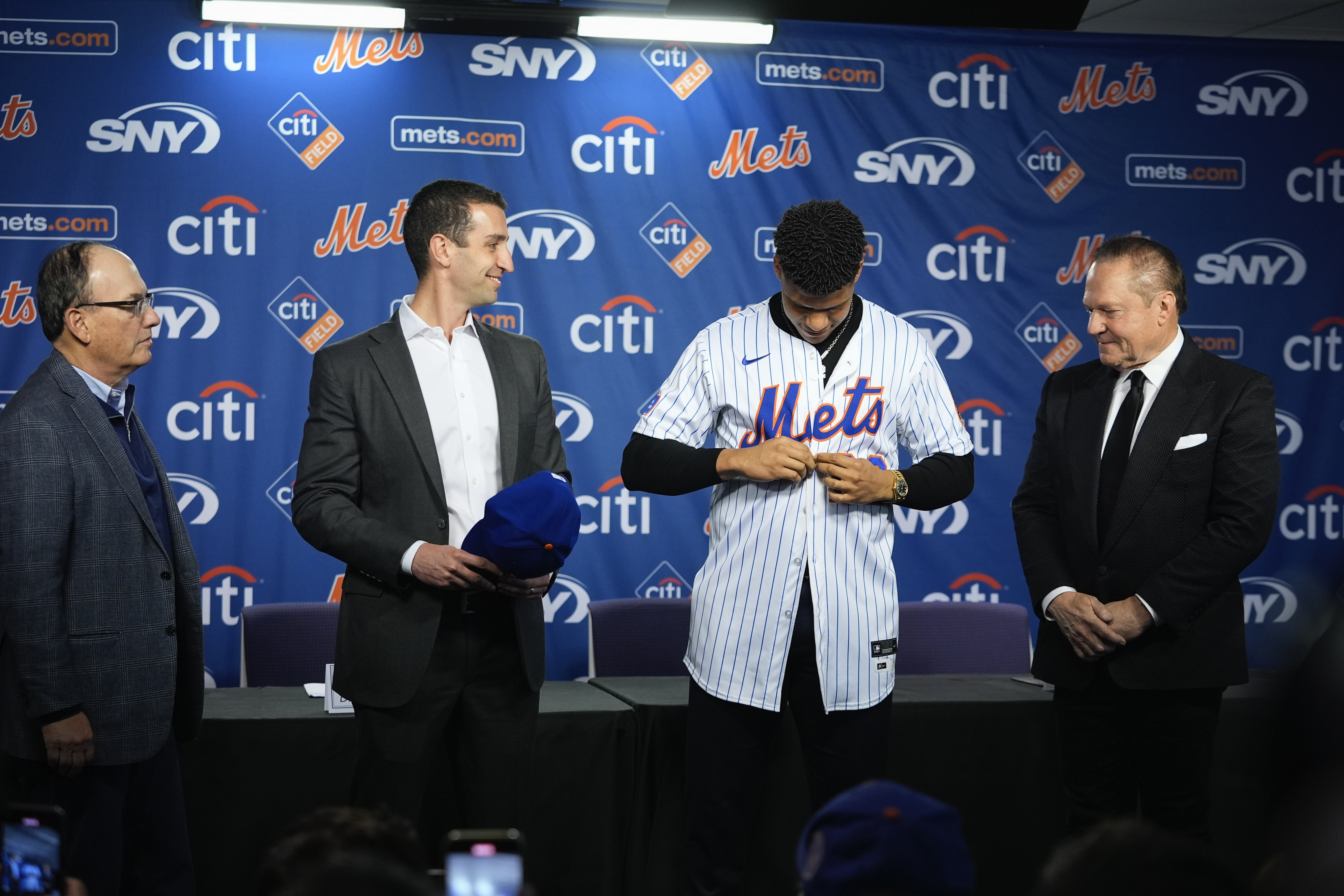 New York Mets' Juan Soto, second from right, puts on a jersey as sports agent Scott Boras, right, Mets owner Steven Cohen, left, and  president of baseball operations David Stearns, second from left, watch during a baseball news conference, Thursday, Dec. 12, 2024, in New York. (AP Photo/Frank Franklin II)