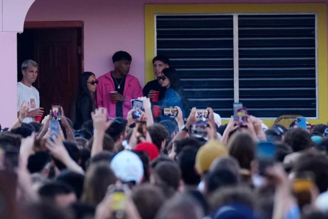 A crowd of people at an outdoor event, with several individuals in the foreground holding cups and phones. Some figures are visible on a balcony or porch in the background.