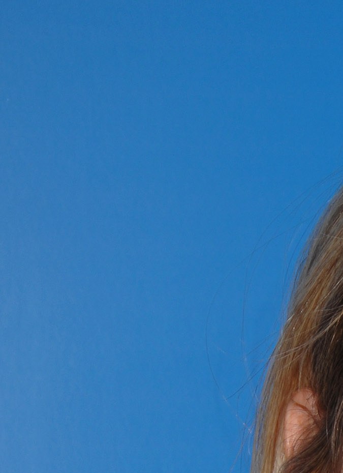 A smiling woman with long, straight blonde hair wearing a white top and a cross necklace, posed in front of a blue backdrop at an event.