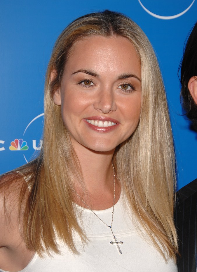 A smiling woman with long, straight blonde hair wearing a white top and a cross necklace, posed in front of a blue backdrop at an event.