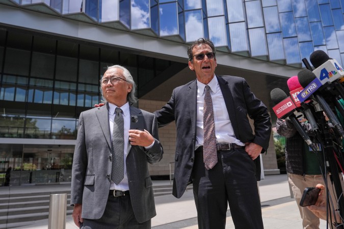 Two men standing outdoors in formal attire, one with glasses and gray hair, and the other gesturing while speaking to a group of reporters. A modern building is visible in the background.
