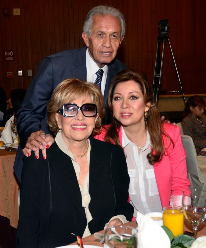 A group of three people smiling for the camera at a dining setting. The foreground features two women, one wearing large sunglasses and a black outfit, and the other in a pink jacket over a white shirt. A man in a suit stands behind them, placing his hand on the shoulder of the woman in black. Table settings with food and drinks are visible.