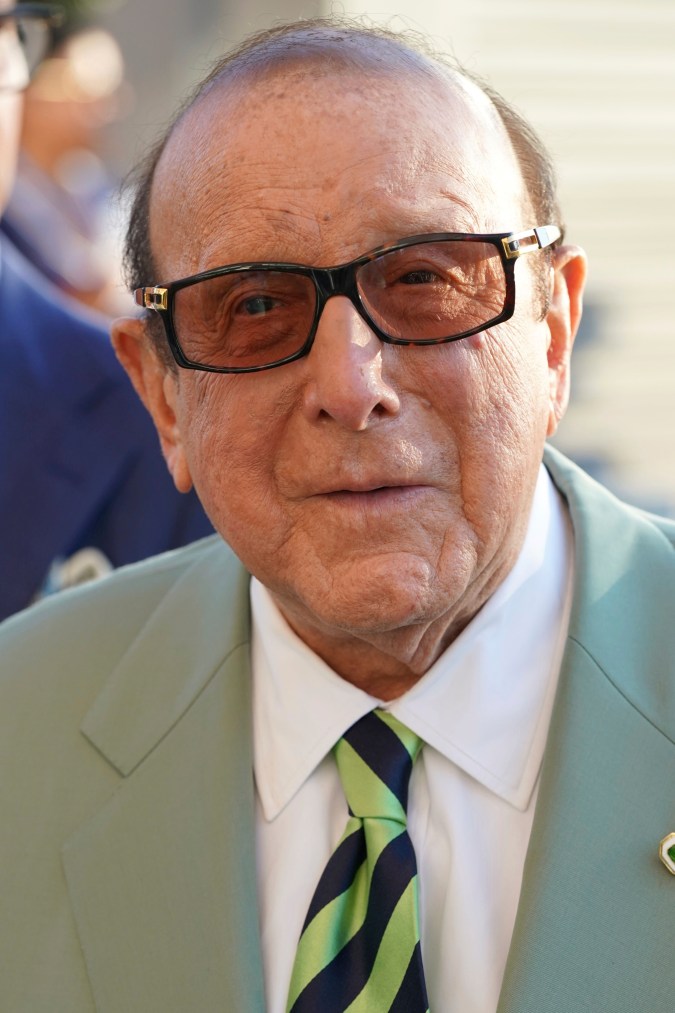 An elderly man wearing sunglasses and a light green suit with a striped tie, smiling at the camera.