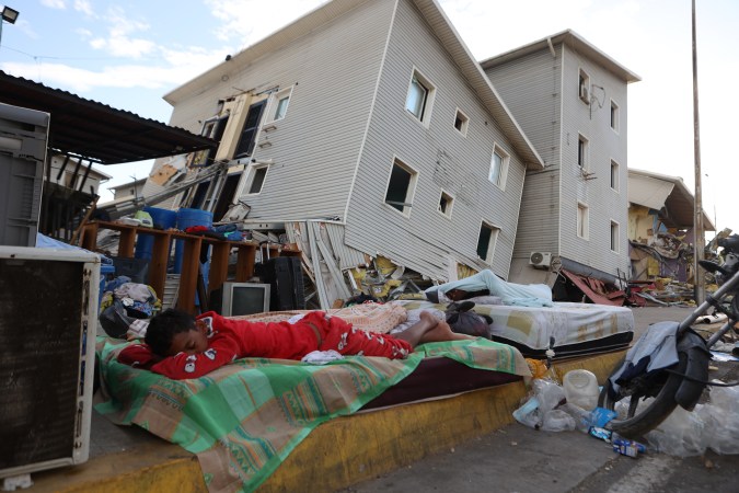 A child sleeping on a blanket in front of a damaged building, with debris and abandoned items scattered around. Several other individuals are resting on mattresses nearby.