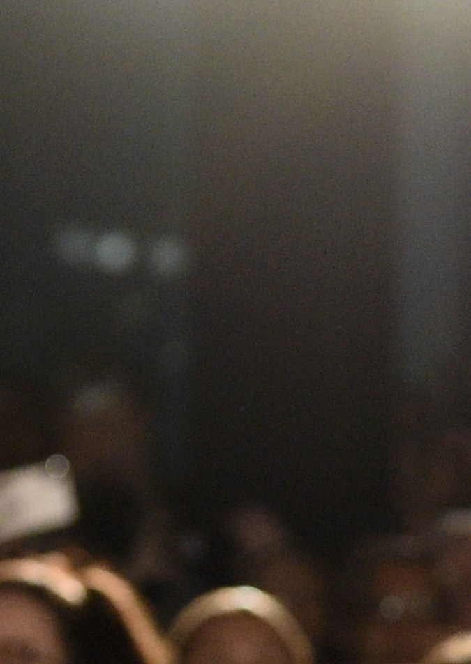 A middle-aged man in a black suit and polka-dot tie is smiling and pointing toward the camera at a film premiere event, with a crowd of fans in the background.