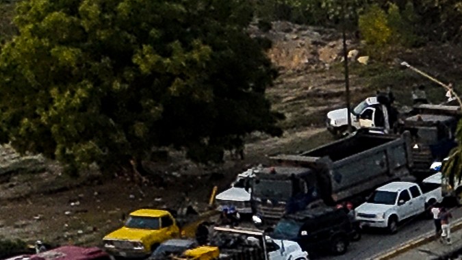 Aerial view of a collapsed building surrounded by debris and damaged structures, with emergency responders at the site and a swimming pool visible nearby.