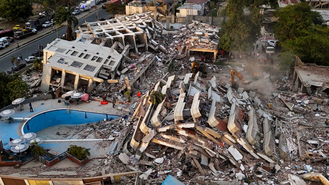 Aerial view of a collapsed building surrounded by debris and damaged structures, with emergency responders at the site and a swimming pool visible nearby.