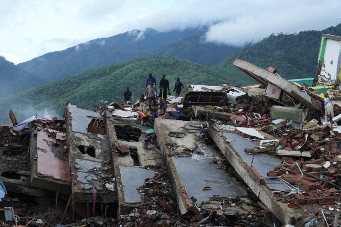 People surveying the debris of collapsed buildings amidst a mountainous landscape after a disaster.
