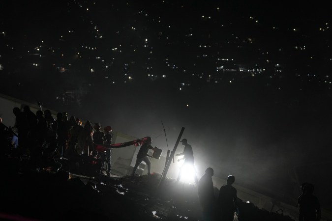 Rescue workers seen silhouetted against a bright light, working amidst debris at night with a backdrop of twinkling city lights.